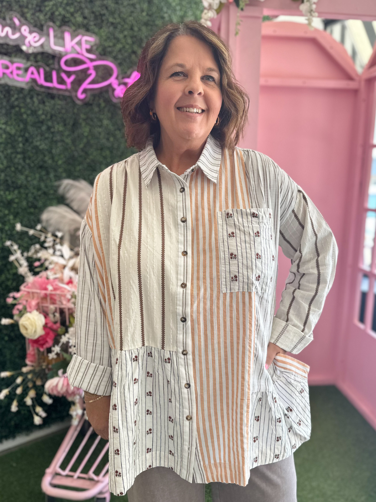 A woman wearing a long-sleeve patchwork button-up shirt with vertical stripes in beige, brown, and white, standing in front of a pink decorative backdrop.