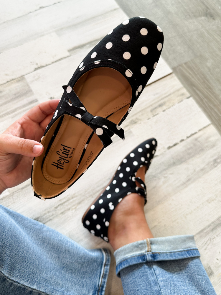 A person holds a black velvet flat shoe with white polka dots and a bow, while wearing the matching shoe on their foot. The brand name &