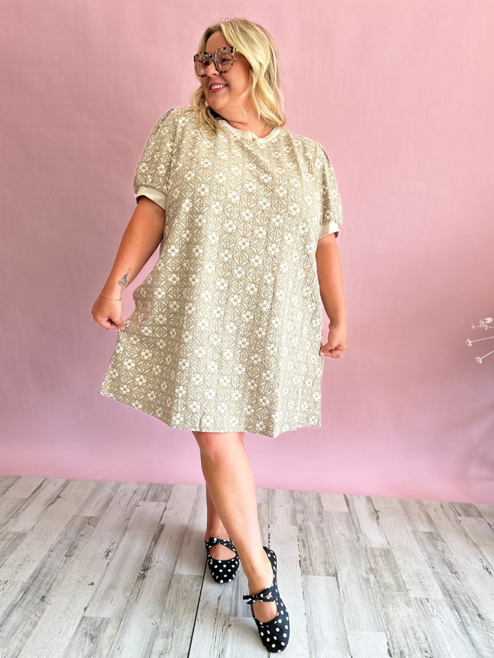 A woman modeling a short-sleeve beige dress with a white floral pattern, standing against a pink studio backdrop with light wood flooring.