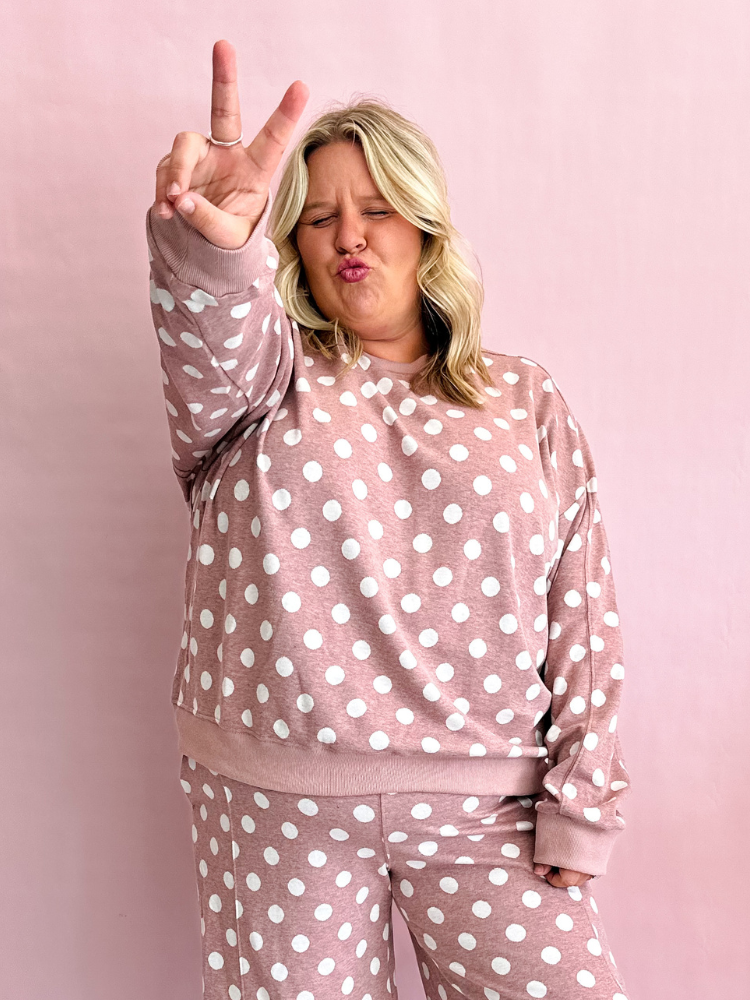 A woman posing in a dusty pink and white polka-dot sweatshirt and matching sweatpants against a solid pink background.