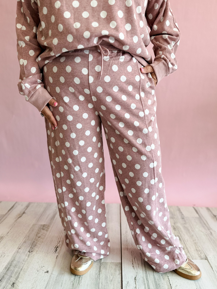A person wearing a dusty rose and white polka-dot lounge set with a matching sweatshirt and wide-leg pants, standing against a pink wall and light wood floor.