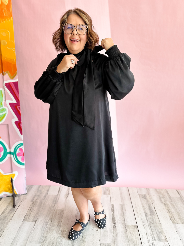 A woman wearing a black satin-like long-sleeve dress with a large bow tie neckline, posing in front of a pink backdrop with a colorful mural on the left.