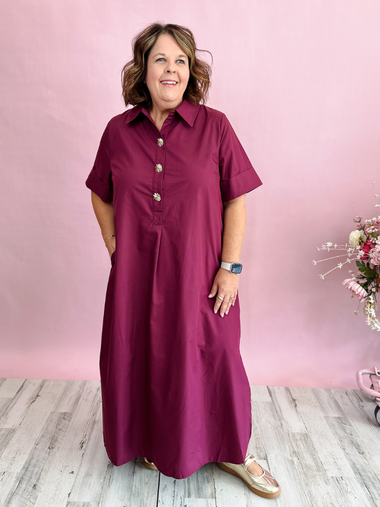 A woman modeling a short-sleeve button-front maxi dress in deep burgundy, standing against a pink wall with light wood flooring.