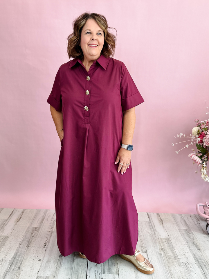 A woman modeling a short-sleeve button-front maxi dress in deep burgundy, standing against a pink wall with light wood flooring.