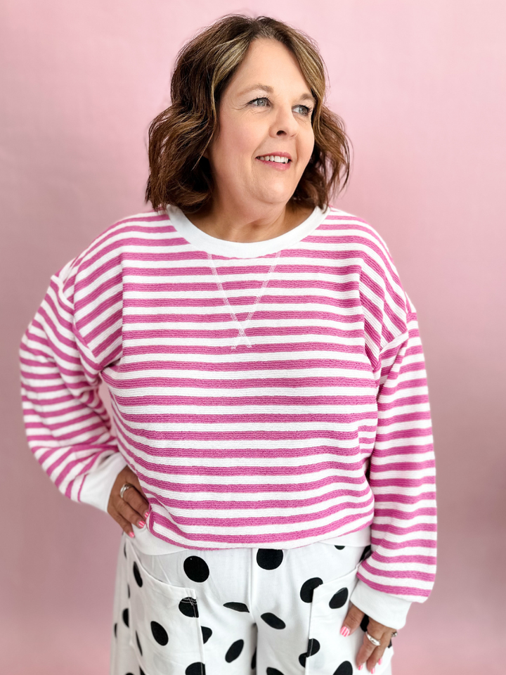 A woman wearing a pink and white horizontally striped long-sleeve crewneck sweatshirt and white pants with black polka dots, standing against a solid pink background.