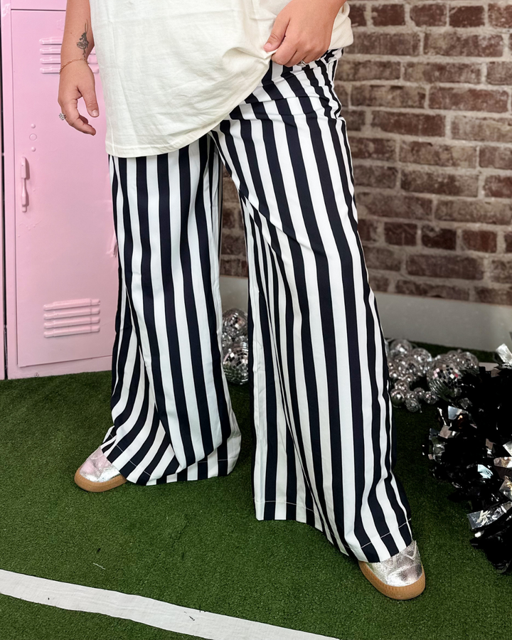 A person wearing black and white vertically striped wide-leg pants, a cream t-shirt, and metallic silver shoes, standing on artificial green turf in front of a pink locker and brick wall.