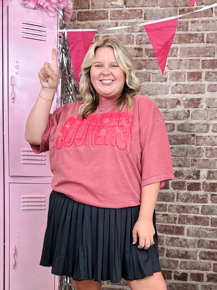 A woman wearing a pink graphic t-shirt and a black pleated skirt stands in front of a brick wall and pink lockers, pointing upward with one finger extended.